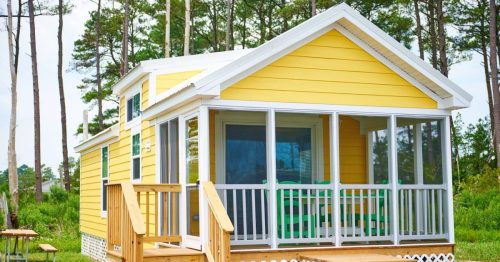 A bright yellow cabin with a screened porch, surrounded by trees and greenery.