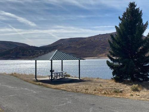 A gazebo by a lake, surrounded by hills and a pine tree, under a clear blue sky.