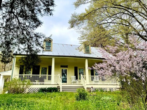 A charming yellow house with a porch, surrounded by greenery and blooming trees under a cloudy sky.