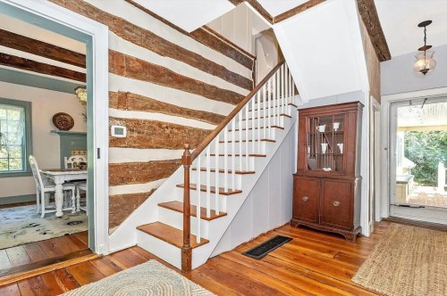 A cozy entryway with wooden beams, a staircase, and a vintage cabinet, leading to a bright outdoor area.