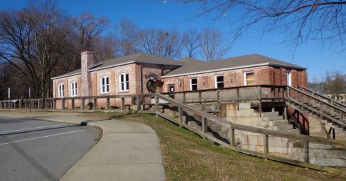 A brick building with large windows, surrounded by trees, featuring a wooden ramp and a paved walkway in front.