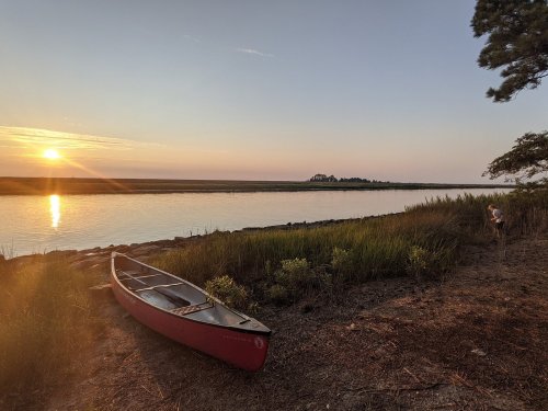 A red canoe rests on the shore at sunset, with calm waters and grassy banks in the background.