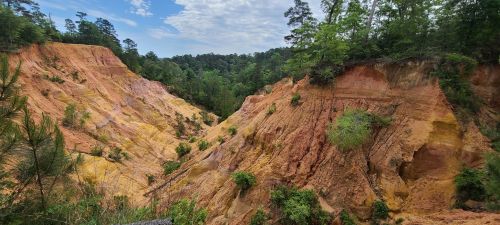 A colorful canyon landscape with steep, eroded walls and lush greenery in the background under a partly cloudy sky.