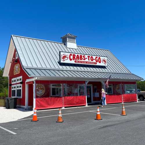 A red seafood market building with a metal roof, featuring a sign that reads "Crabs-To-Go" and a person outside.