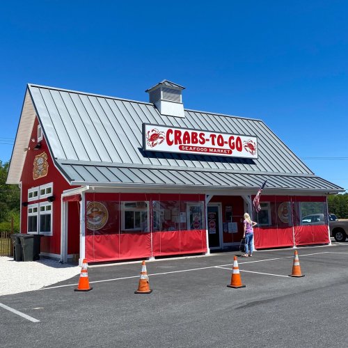 A red seafood market building with a metal roof, featuring a sign that reads "Crabs-To-Go" and a person outside.