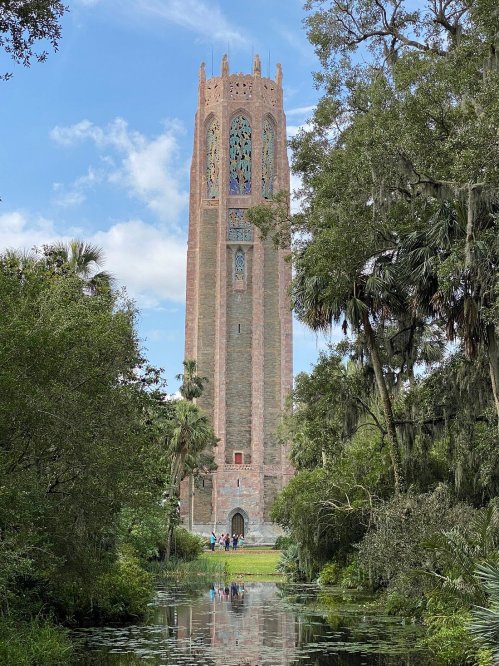 A tall, ornate tower surrounded by lush greenery and a reflective pond under a blue sky with scattered clouds.