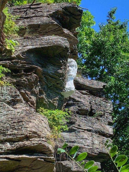 A white rock formation resembling a face on a rugged cliff, surrounded by greenery and blue sky.