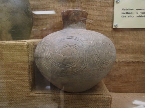 A round, intricately designed pottery vessel with spiral patterns, displayed in a museum case.