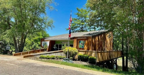 A wooden building with a porch, surrounded by trees, featuring an American flag and colorful decorations.