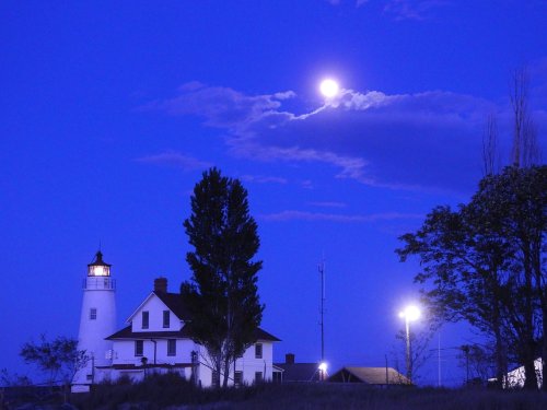 A lighthouse and a historic house illuminated by moonlight under a deep blue sky with clouds.