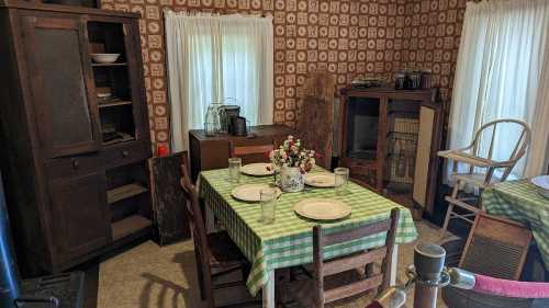 A rustic kitchen with a checkered tablecloth, wooden furniture, and vintage decor, featuring a flower vase and glassware.