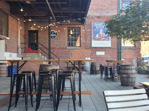 Outdoor seating area with wooden tables, metal chairs, and string lights, set against a brick wall and a brewery sign.