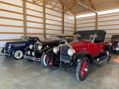 A row of vintage cars in a barn, featuring classic models with shiny exteriors and distinct colors.