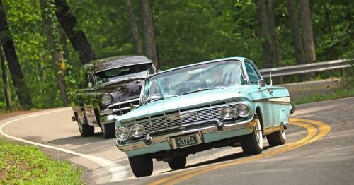 Two classic cars, one turquoise and one black, drive along a winding road surrounded by lush green trees.