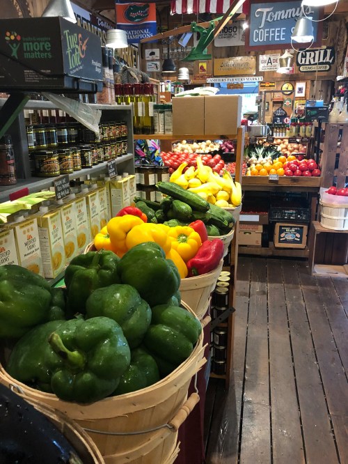 A vibrant market aisle filled with fresh vegetables, including bell peppers, squash, and tomatoes, in wooden baskets.