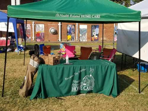 A green tent with a table displaying "Health Related Home Care" in a grassy area, surrounded by colorful chairs.
