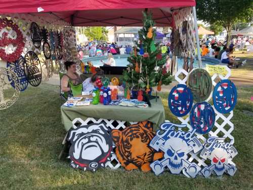 A vendor booth at an outdoor event featuring colorful crafts, decorations, and a woman sitting behind a table.