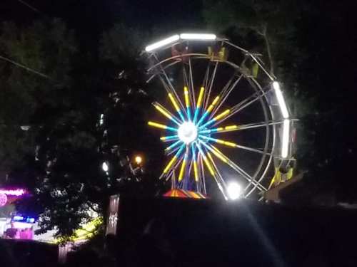 A brightly lit ferris wheel at night, surrounded by trees and colorful lights from a nearby fair.