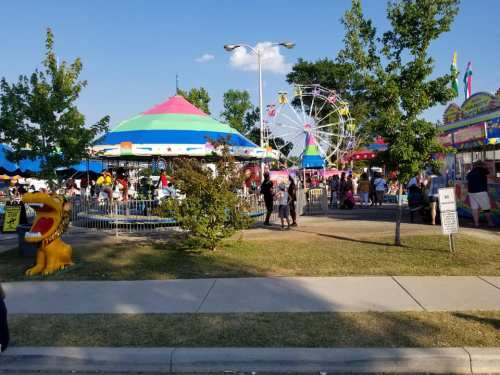 A vibrant carnival scene with a colorful carousel, Ferris wheel, and crowds enjoying various attractions under a clear blue sky.