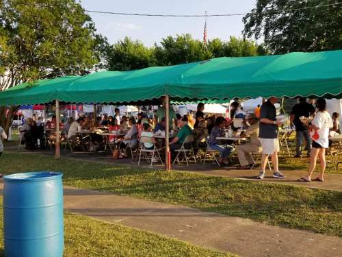 A large group of people dining under a green tent at an outdoor event, with tables set up and a blue barrel nearby.