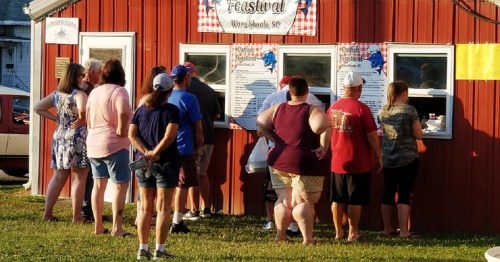 A group of people stands in line at a food stall with a red wooden exterior, waiting to order during a festival.
