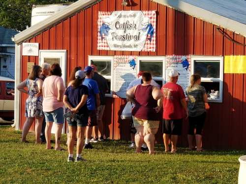 A group of people gathers in front of a booth at the Catfish Festival, looking at the menu.