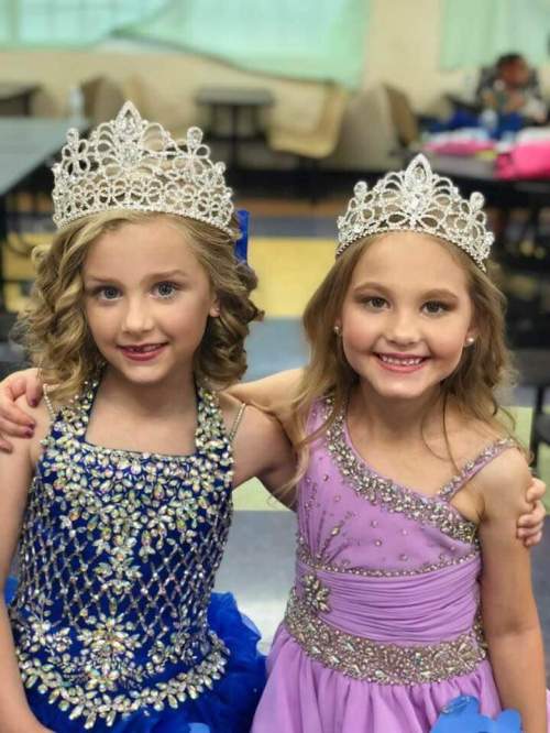 Two young girls in colorful pageant dresses and tiaras smile together, posing happily in a bright indoor setting.
