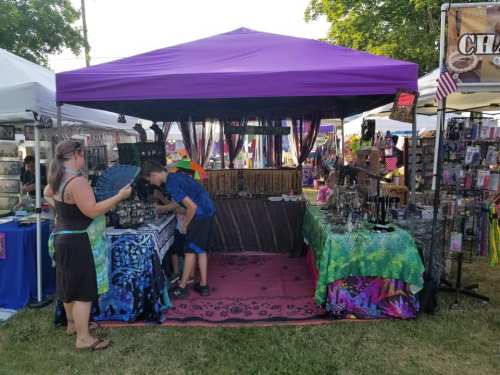 A colorful market stall with jewelry and crafts, featuring a purple canopy and people browsing the items.