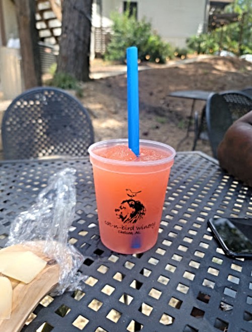 A pink drink with a blue straw sits on a table, next to a snack and a phone, in a sunny outdoor setting.