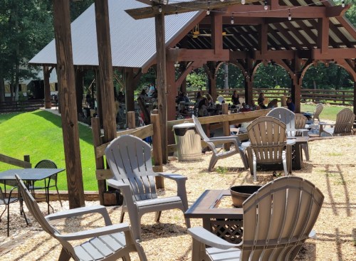Outdoor seating area with wooden chairs and tables under a pavilion, surrounded by greenery and people enjoying the space.