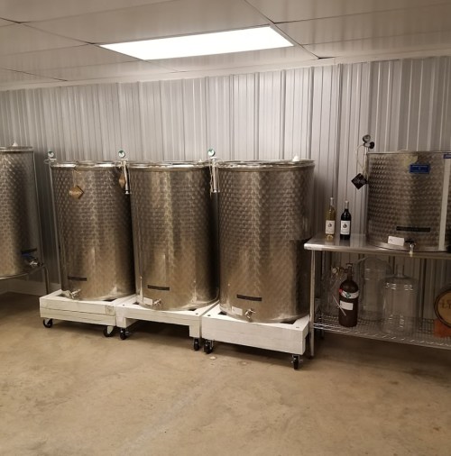 A room with stainless steel fermentation tanks and a metal shelf with bottles and glass containers.