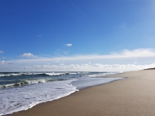 A serene beach scene with gentle waves lapping at the shore under a clear blue sky with scattered clouds.