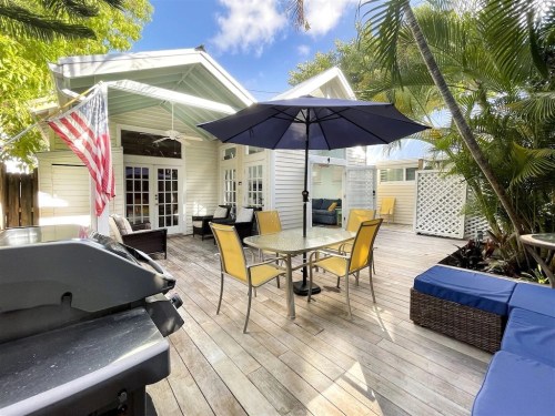 A sunny outdoor patio with a table, chairs, umbrella, grill, and lush greenery, featuring a white house in the background.
