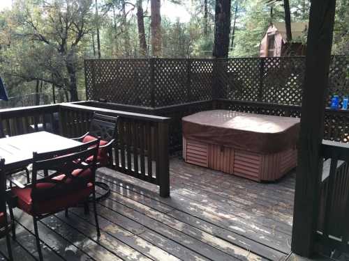 A wooden deck with a hot tub, dining table, and chairs, surrounded by trees and a lattice fence.