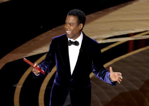 Chris Rock smiles while presenting on stage at an awards ceremony, wearing a velvet suit and holding an envelope.