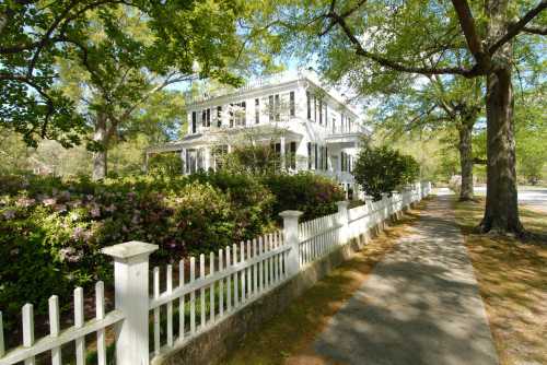 A charming white house with a porch, surrounded by lush greenery and flowers, alongside a tree-lined sidewalk.