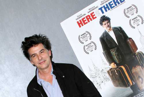 A man with dark, tousled hair stands next to a movie poster featuring him with luggage, promoting the film "Here and There."