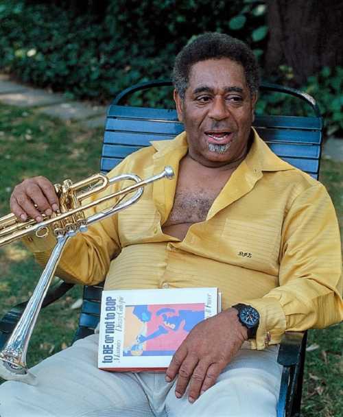 A man in a yellow shirt sits in a chair, holding a trumpet and a book, smiling outdoors.