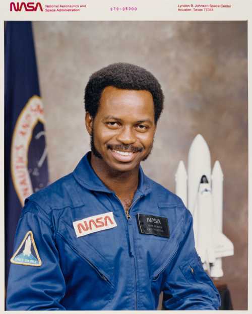 A smiling astronaut in a blue NASA jumpsuit stands in front of a space shuttle model and NASA logo backdrop.