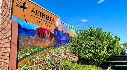 Colorful mural depicting a train and flowers, with "ARTFIELDS" and "LAKE CITY SC" text against a blue sky.