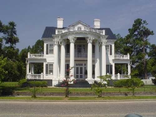 A grand white mansion with columns, a black roof, and lush greenery in front, set against a clear blue sky.