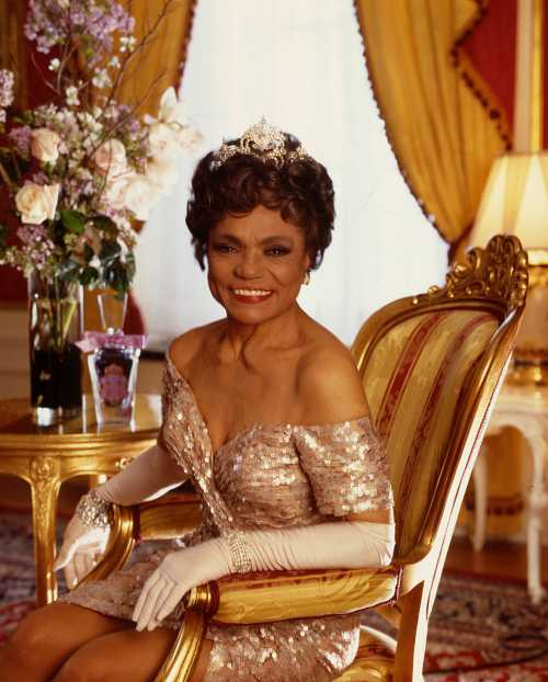 A woman in a sparkling gown and tiara sits elegantly in a lavish room, surrounded by flowers and ornate decor.