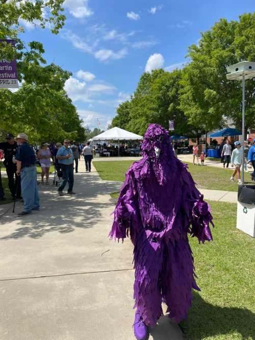 A person in a purple, feather-like costume walks through a festival with tents and crowds in the background.