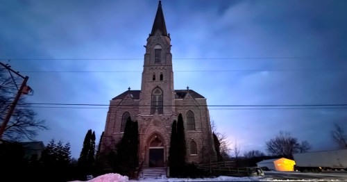 The front facade of a historic stone church in Charlesburg, Wisconsin, featuring a tall steeple and Gothic architecture set against a cloudy twilight sky.