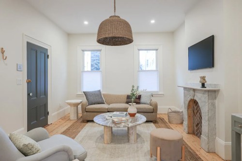 Cozy living room with a light sofa, round coffee table, and a marble fireplace, featuring natural light from two windows.
