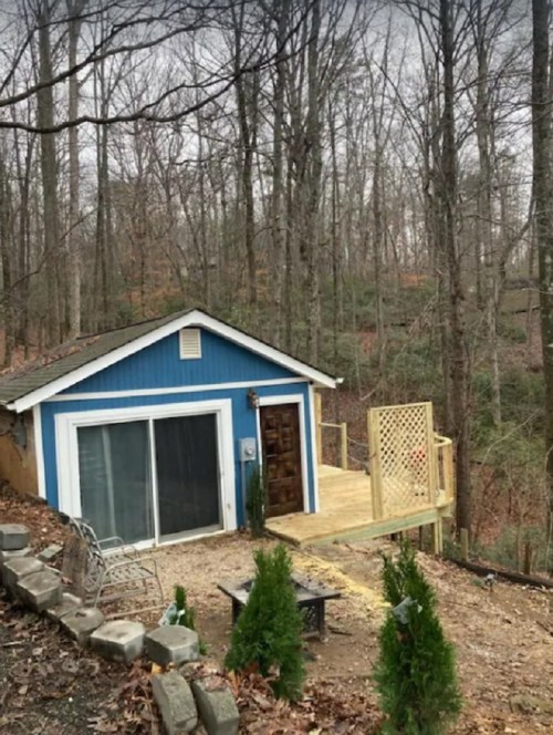 A small blue cabin with a wooden deck, surrounded by trees and a gravel path, in a wooded area.
