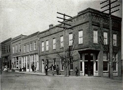 Historic street view featuring brick buildings, storefronts, and people on a quiet road with utility poles.