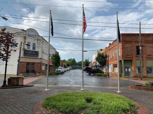 A street view featuring flagpoles, a circular garden, and buildings after rain, with cloudy skies above.