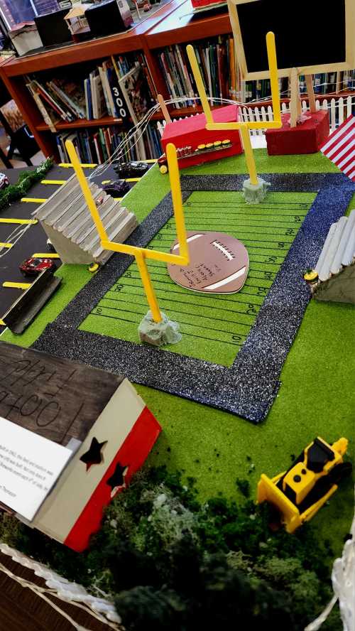 A miniature football field model with goalposts, cars, and decorative elements, set in a library.