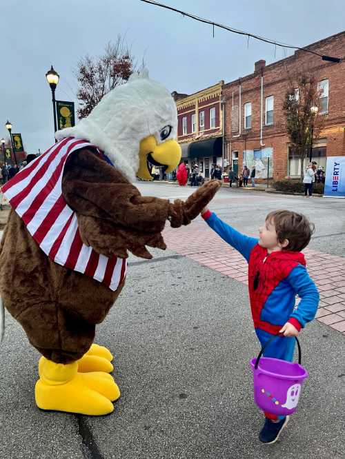 A child in a Spider-Man costume high-fives a large eagle mascot during a festive event on a city street.